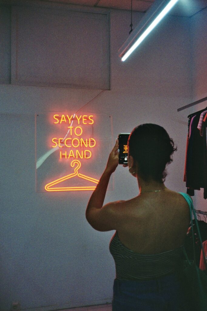 Image shows woman from the back taking photo of a neon sign that reads "Say yes to secondhand"